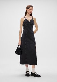 Vestido maxi de ganga preto com alças finas, decote em V e rebites metálicos. Apresenta uma fenda lateral e um design ajustado. Modelo segura uma pequena bolsa preta.