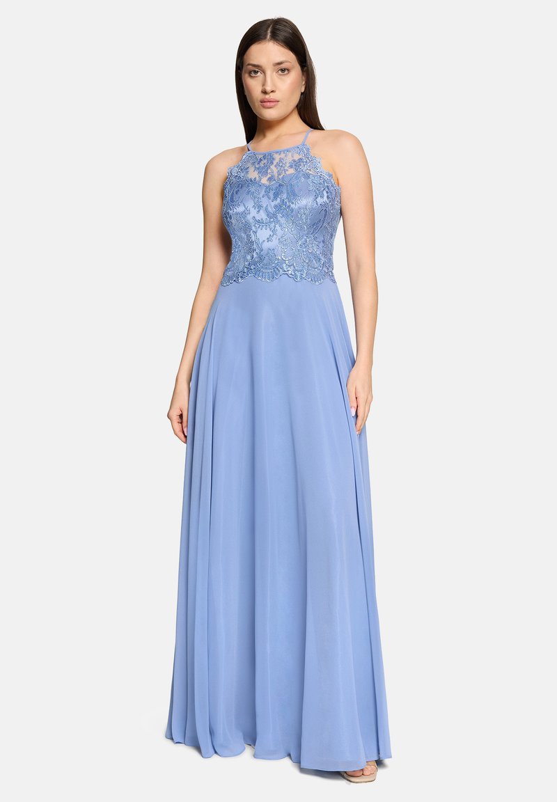 Kobieta w długiej do ziemi, bez rękawów sukni w kolorze niebieskiego dzwonka z koronkowym gorsetem i lejącą się spódnicą z szyfonu, stojąca na tle jednolitego białego tła.