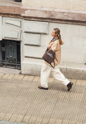 Brązowy plecak z materiału z zamkiem błyskawicznym, noszony na jednym ramieniu. Model w beżowej kurtce i kremowych spodniach, idący po wzorzystym chodniku.