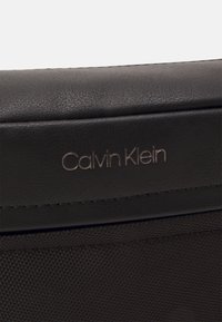 Borsa in pelle nera con superficie liscia e cuciture sottili. Il logo in metallo impresso "Calvin Klein" è ben visibile.