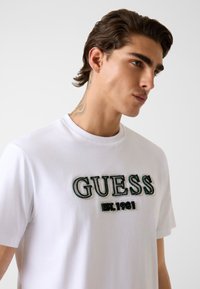 Biała bawełniana koszulka z haftowanym logo "GUESS" w kolorach zielonym i czarnym oraz napisem "EST. 1981" poniżej. Krótkie rękawy i okrągły dekolt.