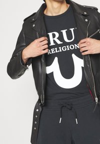 Veste en cuir noir de motard portée sur un t-shirt sombre avec un grand texte blanc "TRUE RELIGION". Le modèle porte un pantalon de survêtement sombre avec cordon de serrage.