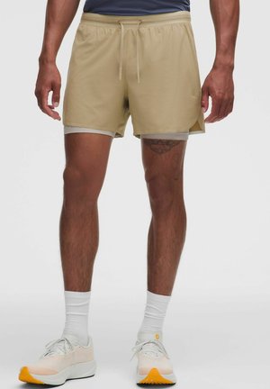 Man draagt beige sportkorte broek met binnenvoering, witte sokken en witte hardloopschoenen met oranje zolen, staand tegen een effen achtergrond.