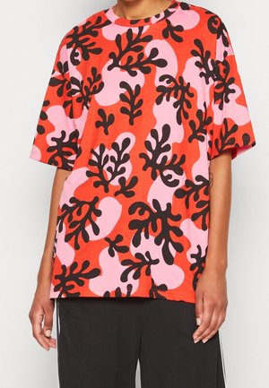 Persoon die een losse korte mouwen T-shirt met rode, roze en zwarte abstracte bladpatronen en zwarte broek draagt.