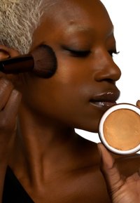 Bronzerkompakt holdes i den ene hånden, påført med en børste i den andre. Mørk hud med en subtil glød, nøytral bakgrunn, minimale detaljer.