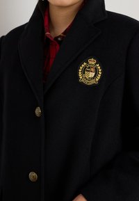 Black wool coat with a notched collar, gold button accents, and a prominent embroidered logo patch on the left chest. Red plaid shirt beneath.