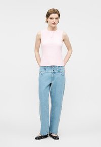 Jeune femme debout, mains derrière le dos, portant un haut sans manches rose clair, un jean taille haute bleu clair et des ballerines noires cloutées.