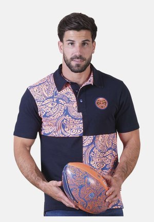 Homme portant un maillot de rugby bleu marine et orange à motifs, tenant un ballon de rugby assorti, faisant face à l'avant sur un fond blanc.