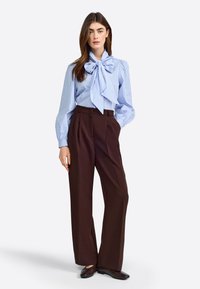 Blau-weiß gestreifte Bluse mit großem Schleifendetail, kombiniert mit hochtaillierten braunen Hosen. Beide Teile verfügen über maßgeschneiderte Schnitte.