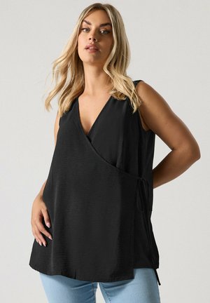 Femme aux cheveux blonds portant un haut noir sans manches à col en V et enroulé, ainsi qu'un jean bleu clair, sur un fond clair uni.