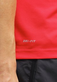 Czerwona koszulka sportowa Dri-FIT o oddychającej fakturze, z srebrnym logo "DRI-FIT" na dolnej stronie. Czarne szorty częściowo widoczne.