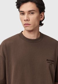 Bruine katoenen t-shirt met een ronde hals en korte mouwen. Heeft een subtiele logo geborduurd op de borst in donkerbruin. Glad textuur.