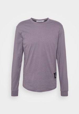 Koszulka z długim rękawem i okrągłym dekoltem w przygaszonym fioletowym kolorze z małą czarną naszywką z logo Calvin Klein blisko dołu po lewej stronie.