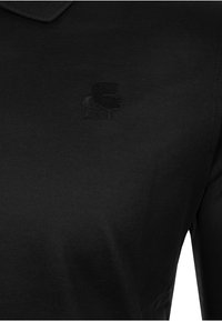 Schwarzes Poloshirt mit einem strukturierten Kragen und gesticktem Logo auf der Brust. Das Material wirkt glatt und leicht.