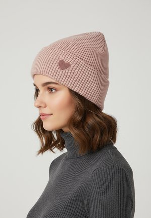Pink knitted beanie with ribbed texture, featuring a small heart emblem on the side. Model wearing a dark gray high-neck sweater.