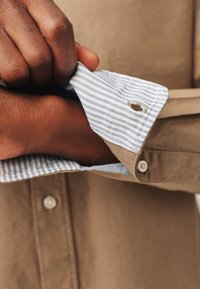 Bruine katoenen shirt met een blauw gestreepte binnenmanchet. De manchet is gedeeltelijk opgerold, waardoor een klein gaatje en een knoopdetail zichtbaar zijn.