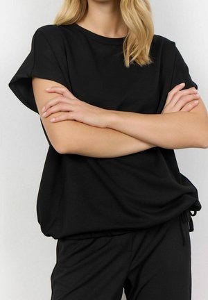 Personne blonde portant un haut noir ample à manches courtes et un pantalon noir assorti, les bras croisés sur la poitrine.