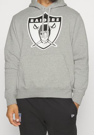 Person trägt einen grauen Raiders Hoodie mit Piratenlogo und schwarze Hose mit NFL-Logo-Patch am linken Bein.