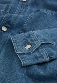 Cămașă din denim cu o culoare albastră clasică, deschidere cu nasturi, nasturi metalici cu capsi, și un guler structurat cu mansete pliate.