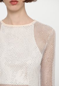 Gjennomsiktig mesh-top med sølvgeometrisk mønster, kremfarget utringning, lange ermer og en lett, teksturert overflate.