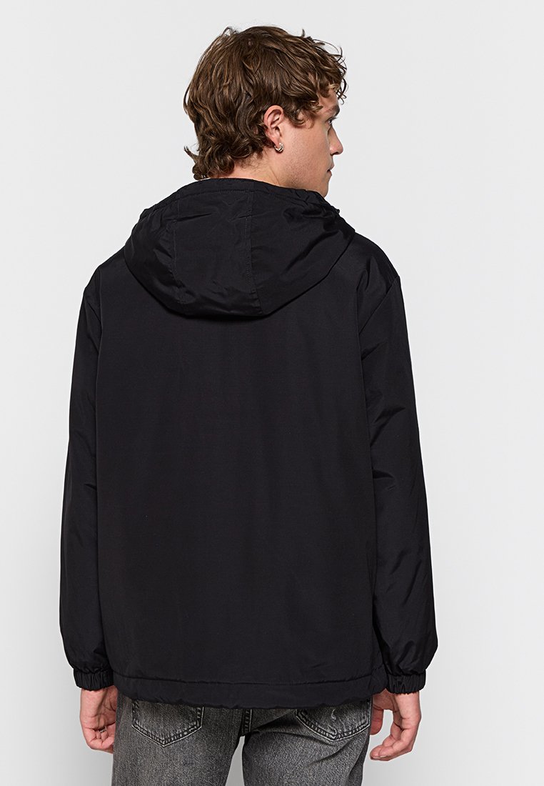 Joven con cabello corto y rizado, vestido con chaqueta con capucha negra y jeans grises, de espaldas frente a un fondo blanco liso.