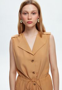 Jumpsuit senza maniche color beige con colletto a punta, bottoni frontali e vita legata. Presenta un tessuto liscio e dettagli di cucitura sottili.