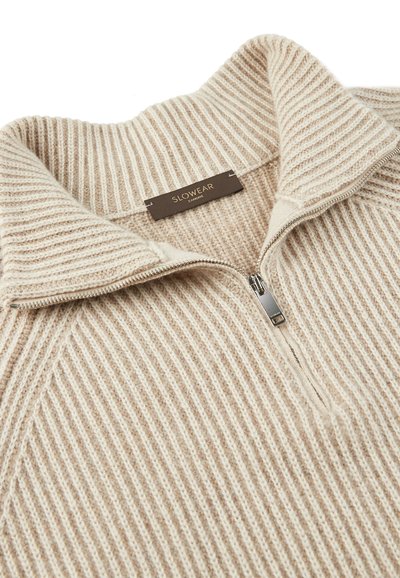 Beige gerippten Pullover mit hohem Kragen und Halbreißverschluss. Die Stofftextur ist weich und gestrickt. Das Markenetikett "SLOWEAR" ist sichtbar im Inneren des Kragens.
