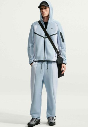 Jeune homme portant un survêtement Nike bleu clair avec capuche relevée, casquette noire, sac bandoulière noir et baskets grises, debout devant un fond uni.