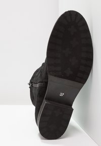 Stivaletto in suede nero con suola in gomma piatta. Presenta un dettaglio con zip e un'etichetta di taglia (37) sulla base. Grip testurizzato sulla suola.