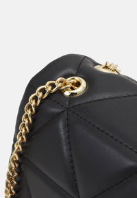 Schwarze Ledertasche mit einem diamantenen Steppmuster, vergoldeten Beschlägen und einem geflochtenen Kettenriemen, der an einer runden metallischen Öse befestigt ist.