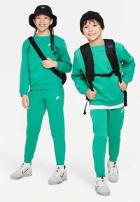 Fatos de treino verdes da Nike para crianças, com ténis a condizer. Tecido suave e corte confortável. Acessórios incluem um chapéu tipo pescador preto e uma mochila.