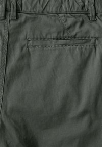 Tmavě zelená látka kalhot, ukazující zadní kapsu s horizontálním otvorem a poutky na pásku.