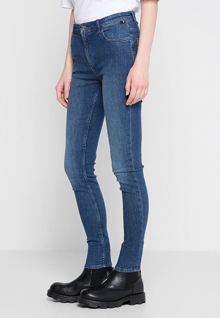 Chude niebieskie dżinsy wykonane z denimu, z średnim stanem, pięcioma kieszeniami i przeszyciami wzdłuż szwów. Noszone z czarnymi botkami za kostkę.