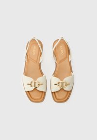 Sandales blanches en simili cuir avec une large sangle et des détails en métal doré. Semelle intérieure beige et design à bout carré. Semelle plate avec une hauteur minimale.