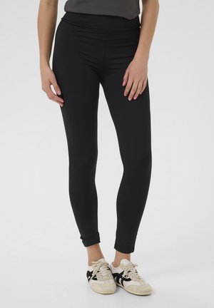 Leggings sportivi neri con vita alta, aderenti, in tessuto liscio e lunghezza alla caviglia, abbinati a sneakers bianche e nere.