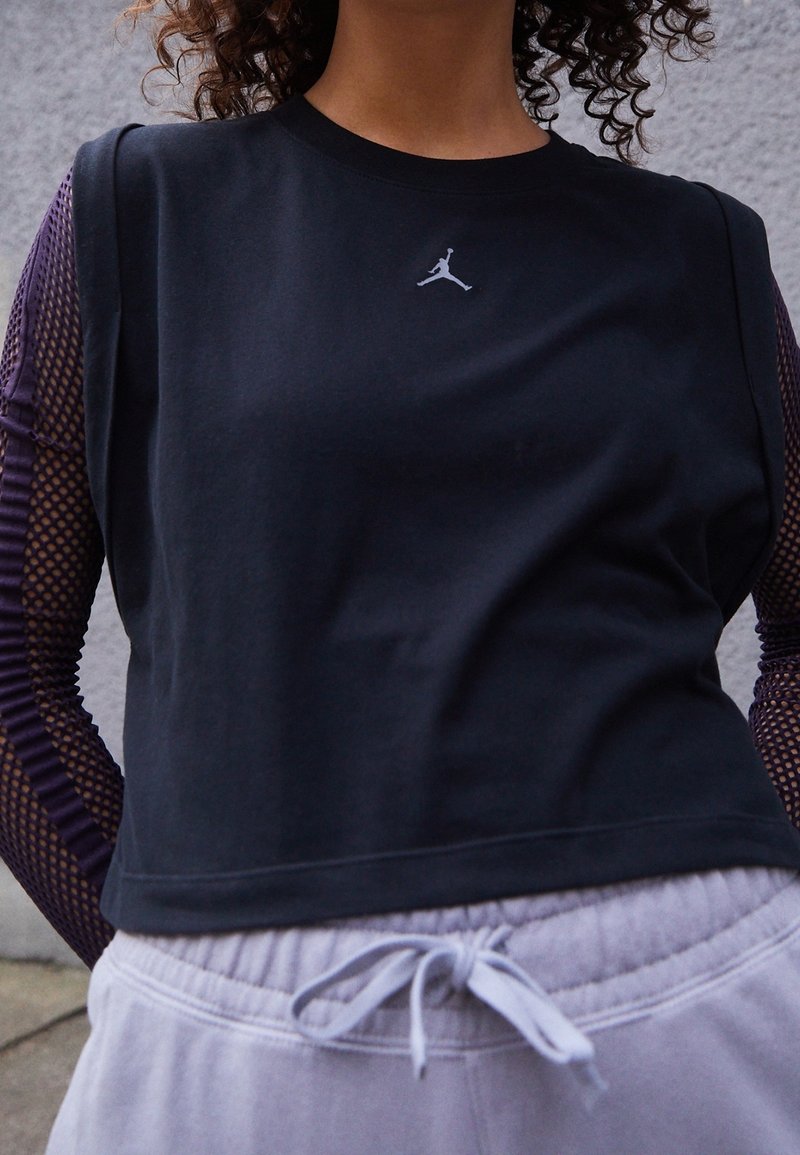 Personne portant un haut noir sans manches avec le logo Jordan sur de longues manches en mesh et un pantalon de survêtement gris clair avec une taille à cordon noué.