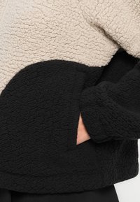 Χέρι που μπαίνει μερικώς στην μπροστινή τσέπη ενός μαύρου και μπεζ υφασμάτινου φλις μπουφάν.