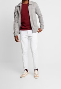 Grå ternet zip-up jakke over en maroon sweater, parret med hvide bukser og beige sneakers med hvide accenter.