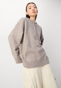 Suéter de punto con capucha de color marrón claro, con un corte relajado, ribete en el dobladillo y mangas largas. Textura suave y diseño minimalista.