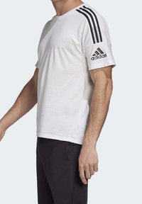 Bílý sportovní tričko s černým designem tří pruhů na ramenou a černým logem Adidas na rukávu. Krátké rukávy a volný střih.