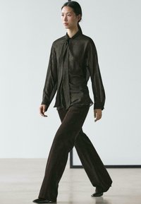 Czarna koszula z długim rękawem z pionowymi złotymi paskami i krawatem, zestawiona z ciemnobrązowymi rozszerzanymi spodniami oraz czarnymi butami, w minimalistycznej scenerii.