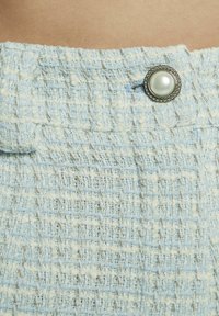 Lichtblauw tweed stof met een gestructureerd patroon. Bevat een ronde parelachtige knop omringd door een zilverkleurige kraalrand.