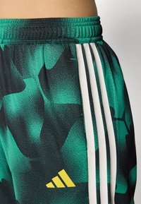Shorts de sport verts et noirs à motifs, comportant trois bandes blanches sur les côtés et un logo jaune sur le bas de la jambe.