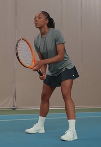 Tenisa spēlētājs pelēkā īsā piedurkņu krekla un melnu šortu tērpā, tur oranžu raketi, ģērbies baltās kurpēs un zeķēs uz zilā laukuma.