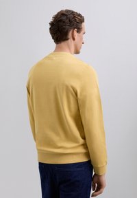 Mann mit kurzem lockigem Haar, der einen gelben Sweatshirt und dunkle blaue Hosen trägt, seitlich vor einem schlichten grauen Hintergrund stehend.