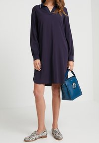 Mörkblå skjortklänning med lång ärm och krage. Buren med mönstrade skor. Accessoarer inkluderar en teal handväska med en cirkulär detalj.