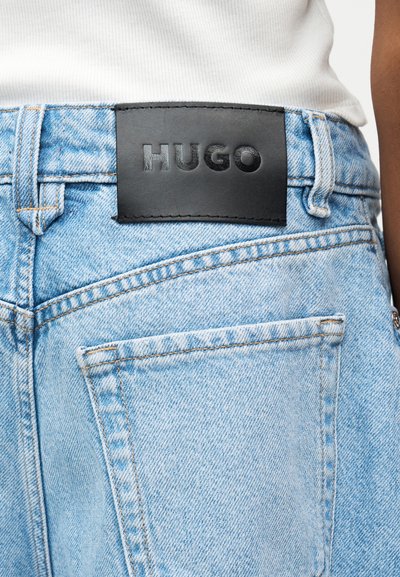 Човек, носещ светлосини дънки от деним с черен кожен етикет с надпис "HUGO" на колана, комбинирани с бял топ.