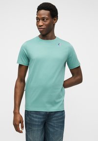 Νέος άνδρας που κοιτάζει πλάγια, φοράει ανοιχτό πράσινο κοντομάνικο πουκάμισο με μικρό λογότυπο και μπλε τζιν, στέκεται μπροστά σε απλό φόντο.