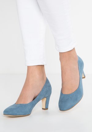 Niebieskie zamszowe szpilki z okrągłym noskiem i krótkim, smukłym obcasem, noszone z białymi spodniami 3/4.
