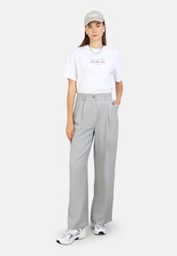 White t-shirt with red text, grey high-waisted trousers, and white sneakers. The outfit is completed with a light grey cap.